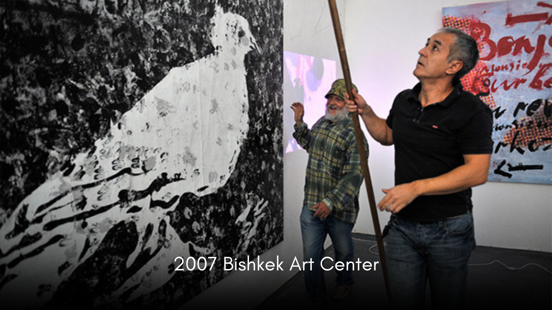Two men standing in front of abstract black and white artwork at Bishkek Art Center in 2007. One man is smiling and gesturing with his hand; the other man is holding a staff and looking upwards.