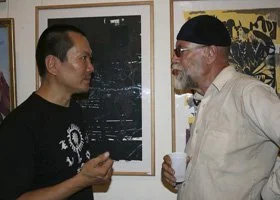 Two men facing each other in a conversation at an art gallery. One man is wearing a black t-shirt, and the other is wearing a white shirt with sunglasses.
