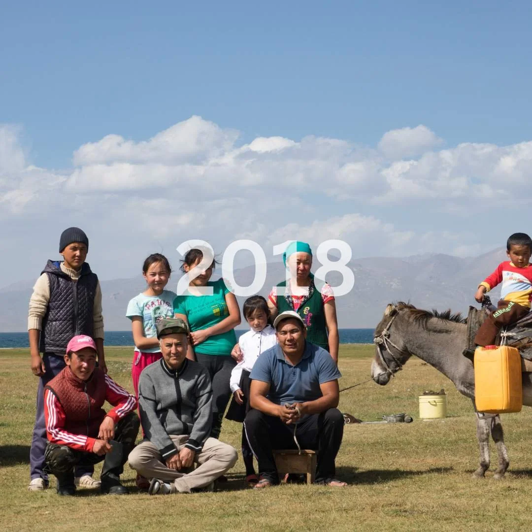 A group of ten people, including children, adults, and an elderly person, posing outdoors on a grassy field with a horse on the right and a mountainous landscape in the background. The image has the year "2018" overlaid at the center.