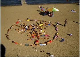 People forming a peace sign on a sandy beach with towels, umbrellas, and individuals relaxing.
