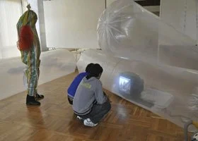 Two people, one standing and one squatting, are observing an object covered in plastic on the floor. The object appears to be a small television or monitor emitting a bright light.