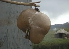 A large clay pot hanging outdoors with a stick, against a cloudy sky and a grassy landscape with a tent in the distance.