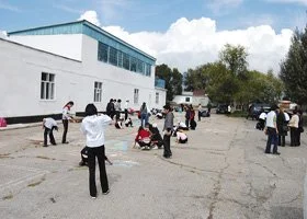 People gathering outside a white building on a sunny day, some wearing hats, with a few cars parked nearby.