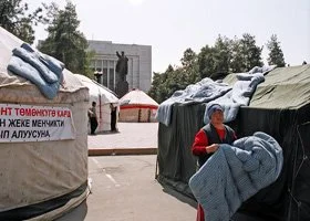 People in a tent village with tents and signs, some wearing hats, outdoors with trees and buildings in the background.