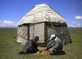 Two people sitting on the grass outside a traditional round yurt under a blue sky.