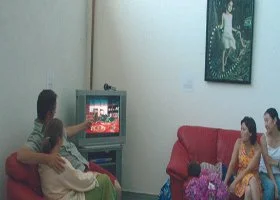 Two women sitting on a sofa watching TV, with one woman hugging a child. Another woman and a child are sitting on a red armchair nearby. The room has a painting on the wall and a bouquet of flowers on a small table.