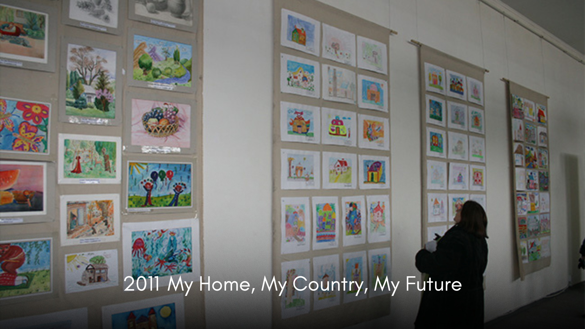 A woman viewing colorful children's artwork displayed on beige fabric-covered display boards in an art exhibition, with the caption "2011 My Home, My Country, My Future" at the bottom.