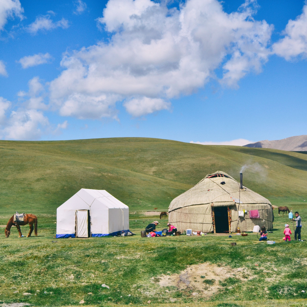 A rural landscape with two tents, one white and one thatched, set on green grass under a blue sky with scattered clouds. People and horses are nearby, with mountains in the distance.