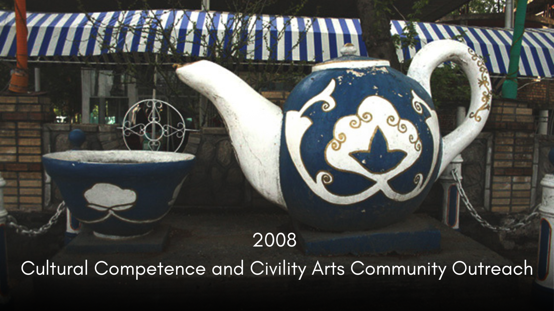 A large decorative teapot with a plus-shaped design on its body, painted in blue and white with gold accents, placed outdoors on a stone platform. The teapot's spout is elongated and curved, and its handle is large and round. Nearby is a small matchi