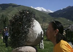 A person leaning in to touch or kiss a stone sculpture with water splashing around it, outdoor setting with mountains in background.