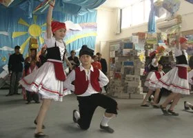 Children performing a traditional dance, dressed in colorful folk costumes, in a classroom or community center setting with decorations and posters in the background.