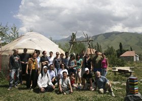Group of people outdoors near a traditional yurt, with a wooden structure and mountains in the background.