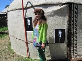 Woman with curly hair wearing colorful clothing standing outside a stone structure with black and white photographs on the wall.