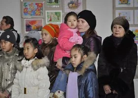 Group of children and two women standing indoors, some children wearing winter clothing, surrounded by colorful artwork on the walls.
