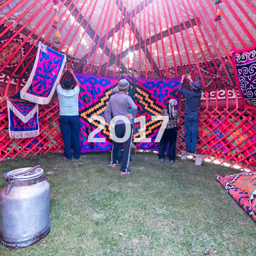 People decorating a colorful, patterned textile inside a traditional yurt with red lattice walls during daylight, with the year 2017 overlayed.