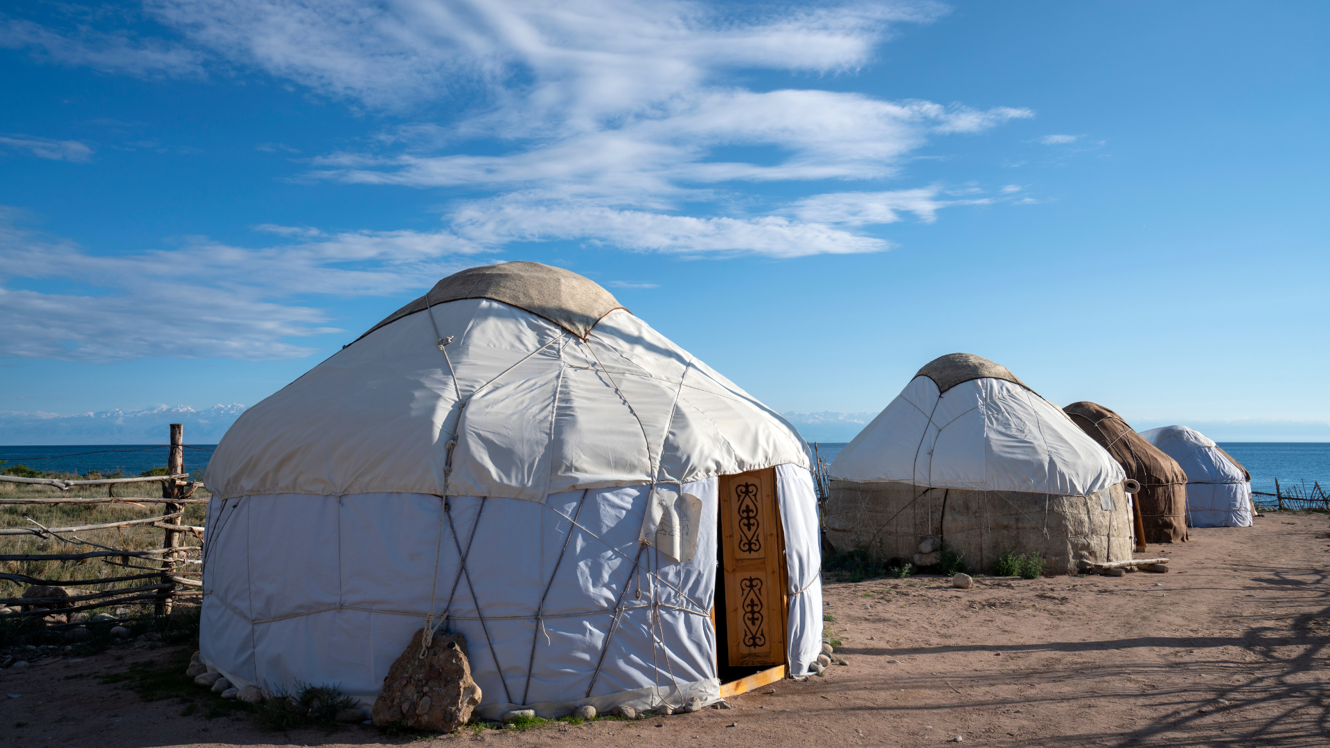 Multiple traditional yurts near the sea under a blue sky with scattered clouds.