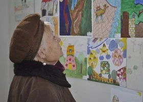 An elderly woman wearing a brown hat and coat looks at children's drawings and paintings displayed on a wall.