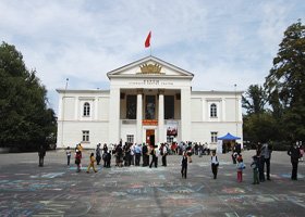 A large white government building with a red flag on top, people gathered in front, and trees on either side.
