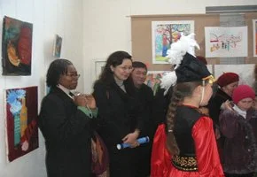 Group of people viewing colorful artwork in an art gallery, including a woman dressed in a Native American costume with a feathered headdress.