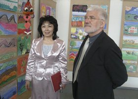 A woman in a shiny silver jacket and a man with white hair and glasses standing in front of colorful artwork displayed on walls.