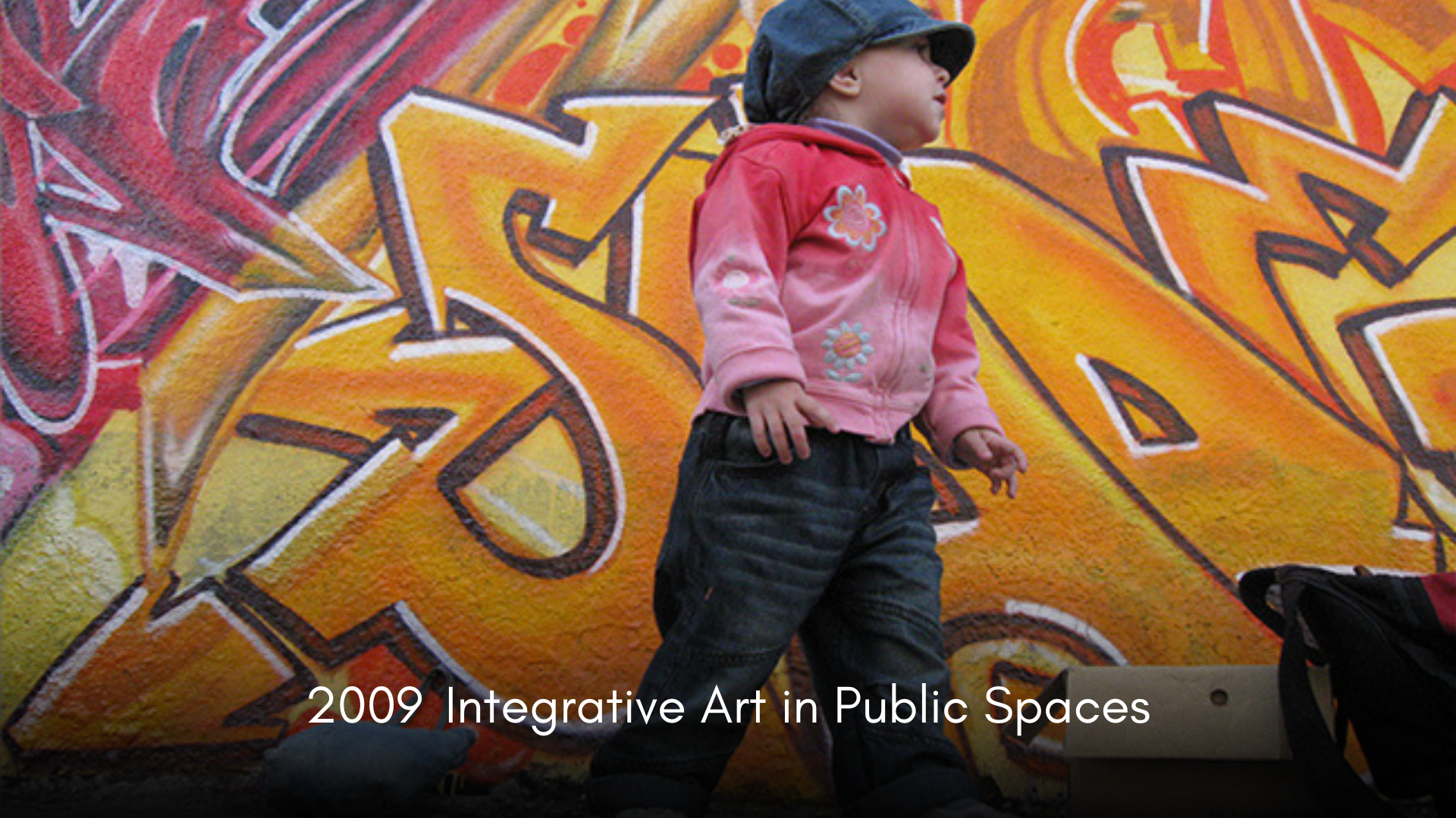 Child wearing a pink jacket with floral embroidery, black hat, and dark pants, standing in front of a colorful graffiti mural in an urban environment.