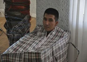 Man with short dark hair, wearing a plaid shirt, sitting at a table with a checked tablecloth, in a room with light-colored curtains.