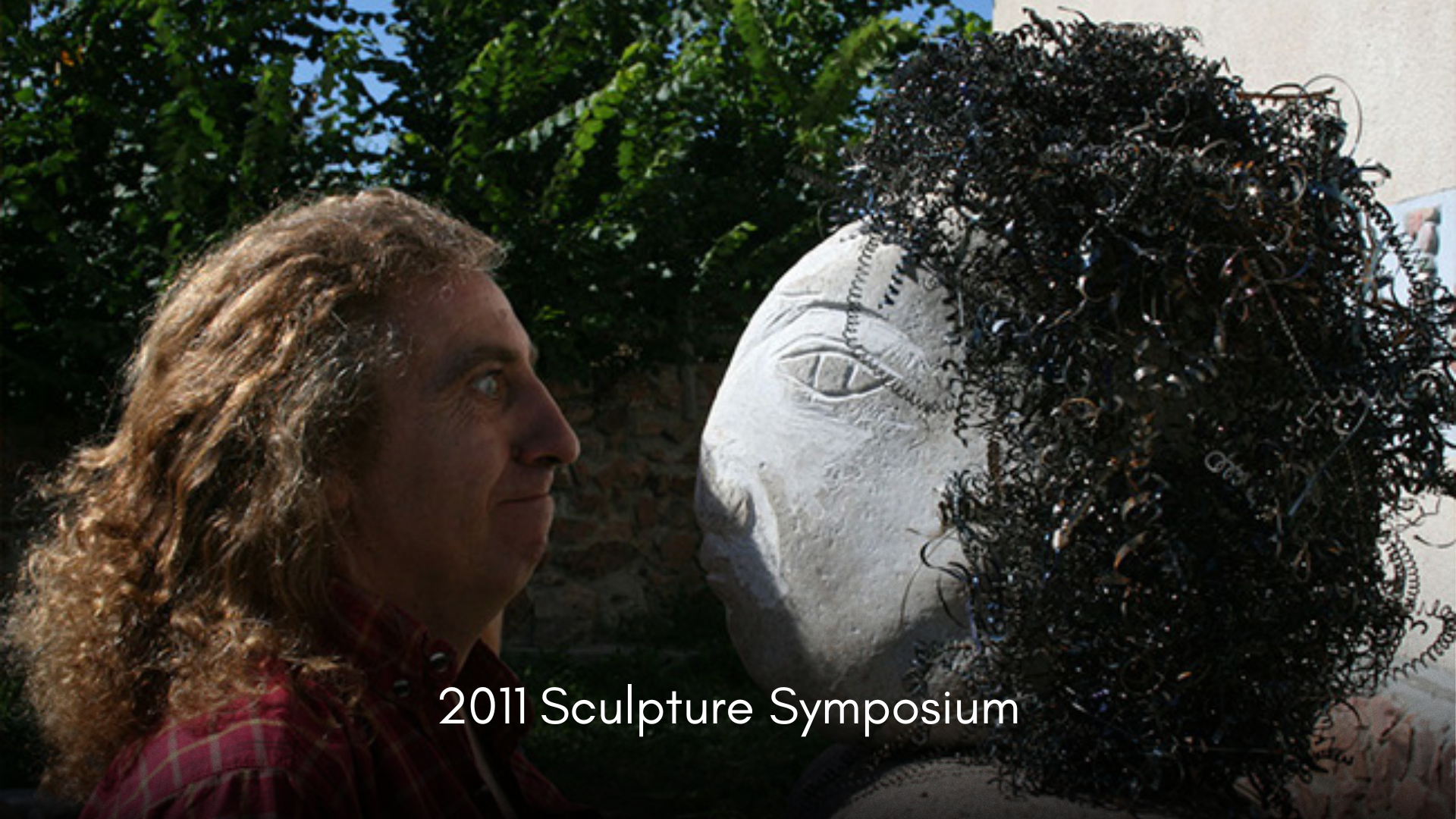 A man with long, curly hair and a red plaid shirt looking at a large sculpture of a woman's face with curly hair made of metal wire, outdoors with green foliage in the background.