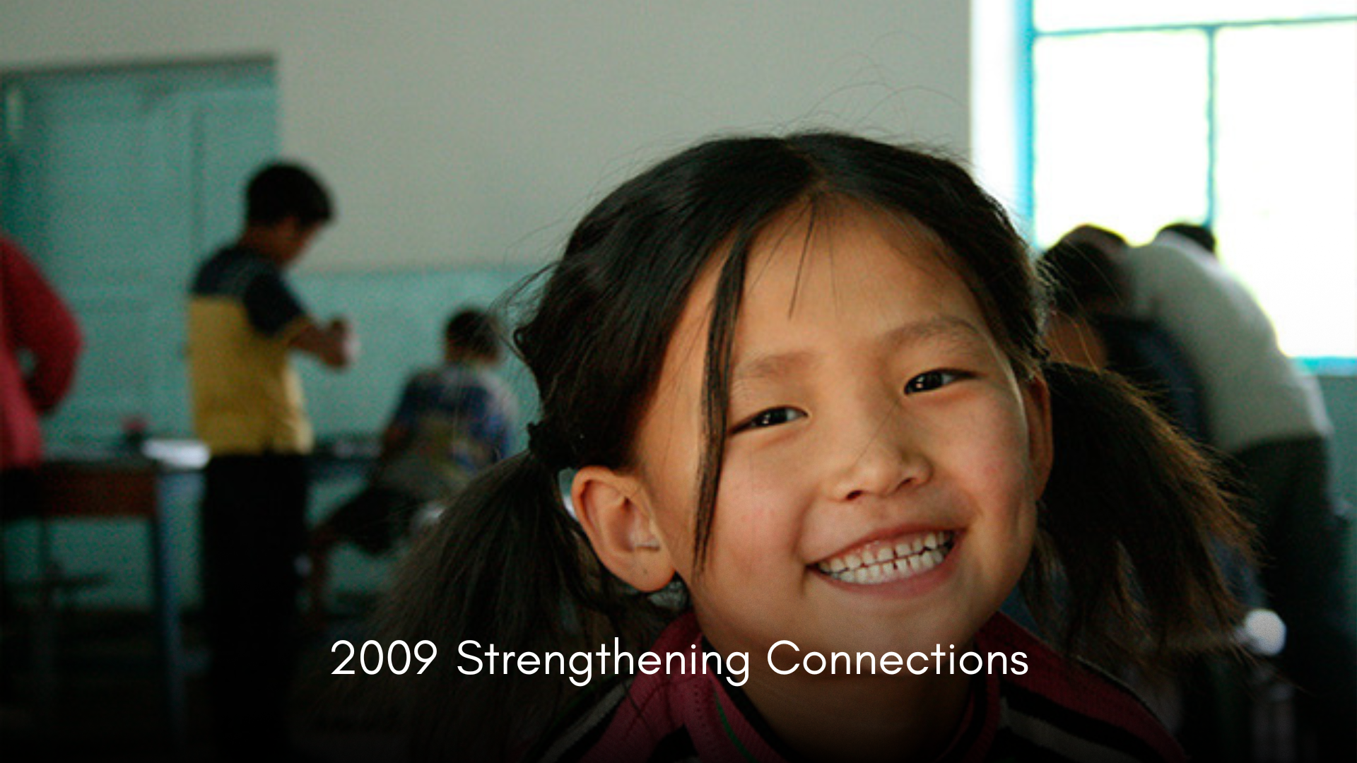 Close-up of a smiling young girl in a classroom, with other students in the background.