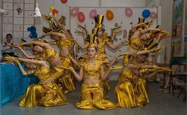 A group of nine young girls dressed in gold costumes performing a traditional dance on a stage decorated with pink and orange balloons.