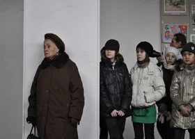 A woman standing alone in front of four children who are lined up and waiting, in an indoor setting with colorful posters on the wall.