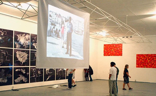 People viewing artwork and a large projected image in an art gallery with black and white images on the wall and red abstract paintings.