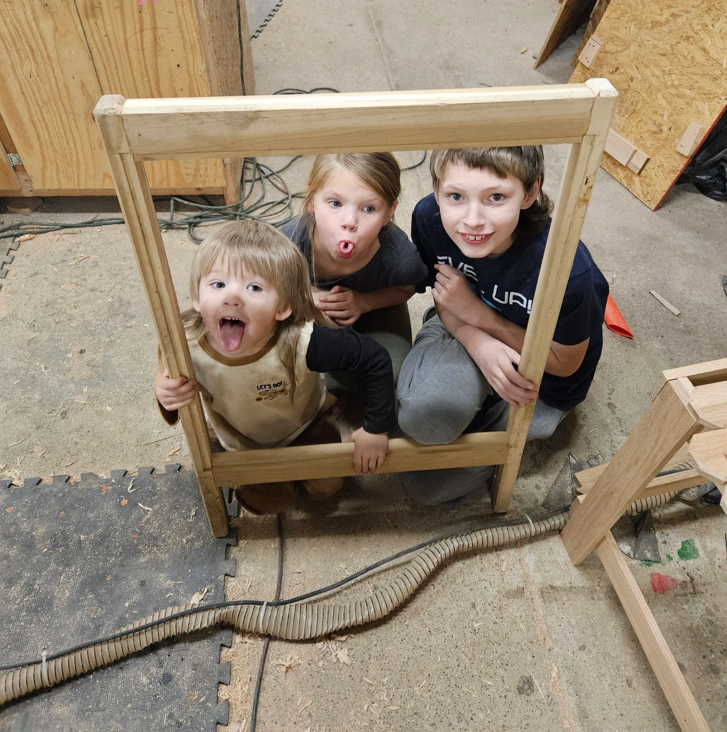 Three children playfully peering through an unfinished wooden frame in a workshop or woodshop.