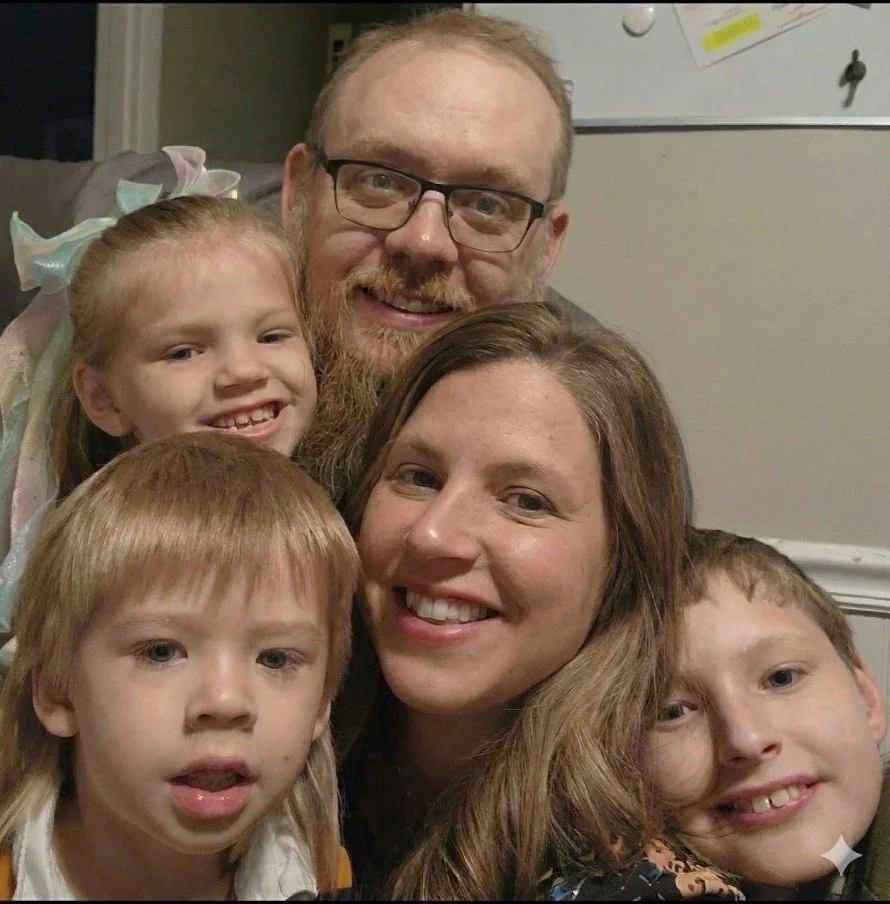 Family of five smiling together, with a man with glasses and a beard, a woman with long brown hair, and three children, two boys and a girl, in a cozy indoor setting.