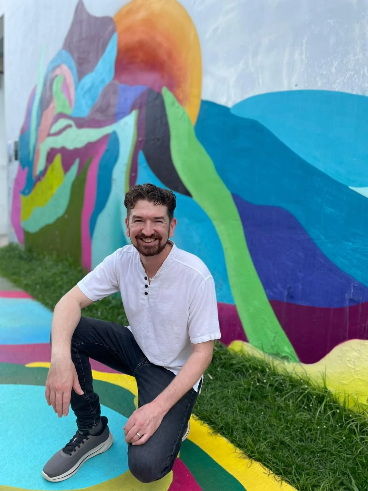 A smiling man with dark curly hair and a beard crouching in front of a colorful mural on a wall, with vibrant abstract shapes and patterns.