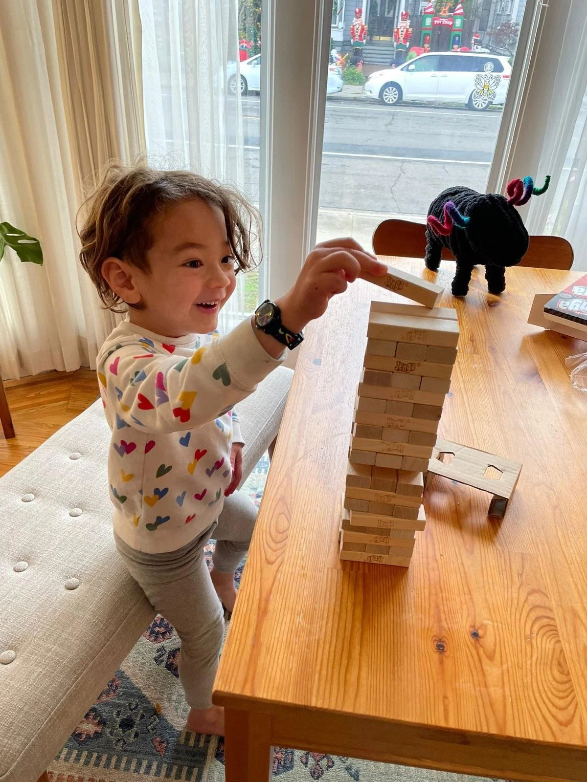 A man sitting on a beige cushioned bench against a cream-colored wall, wearing a blue short-sleeved sweater and beige pants, with slicked-back dark hair and a calm expression as he looks to the side.
