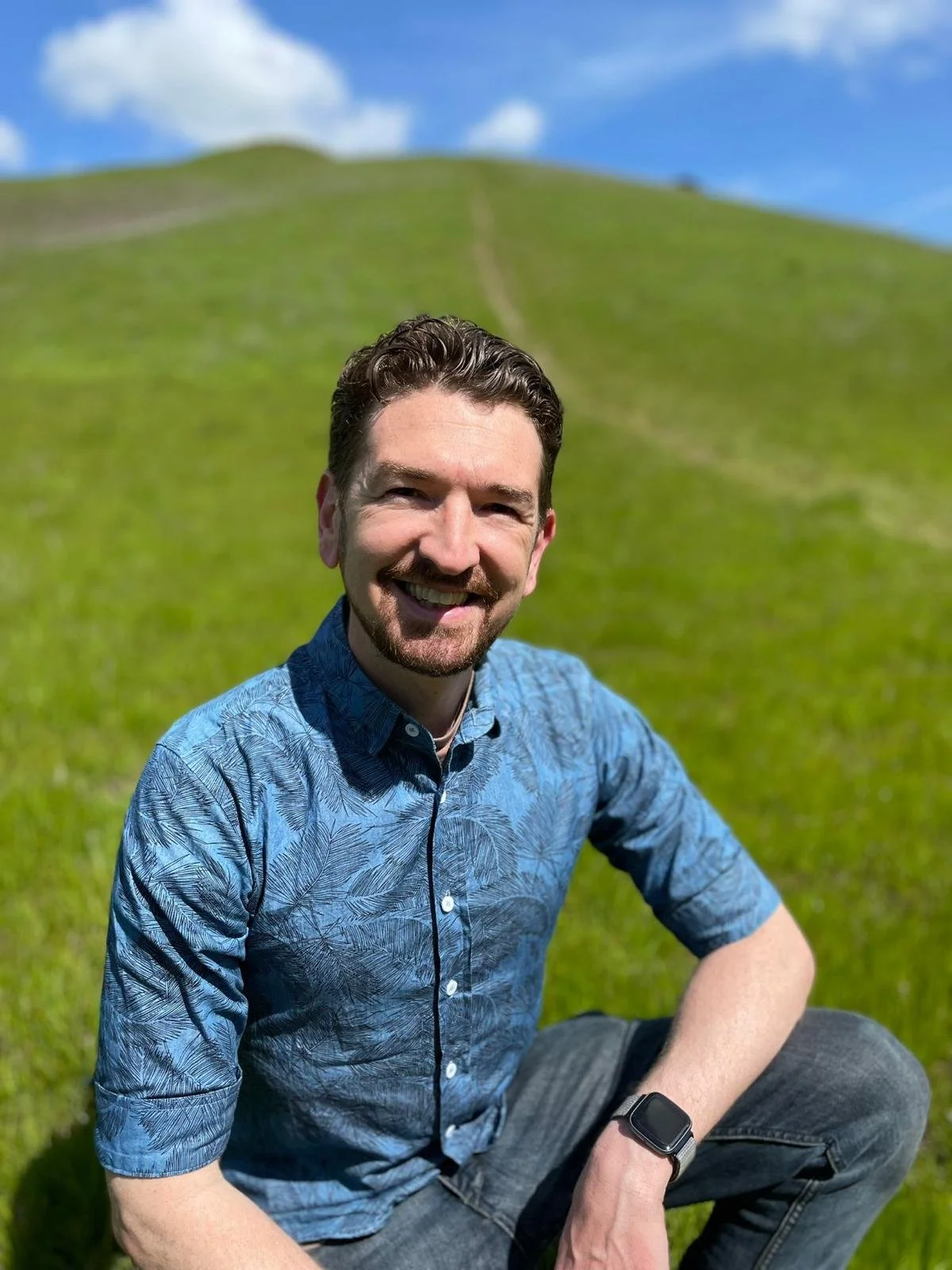 A smiling man with a beard, wearing a blue patterned button-up shirt and a smartwatch, sitting on grass with a green hill and blue sky with clouds in the background.