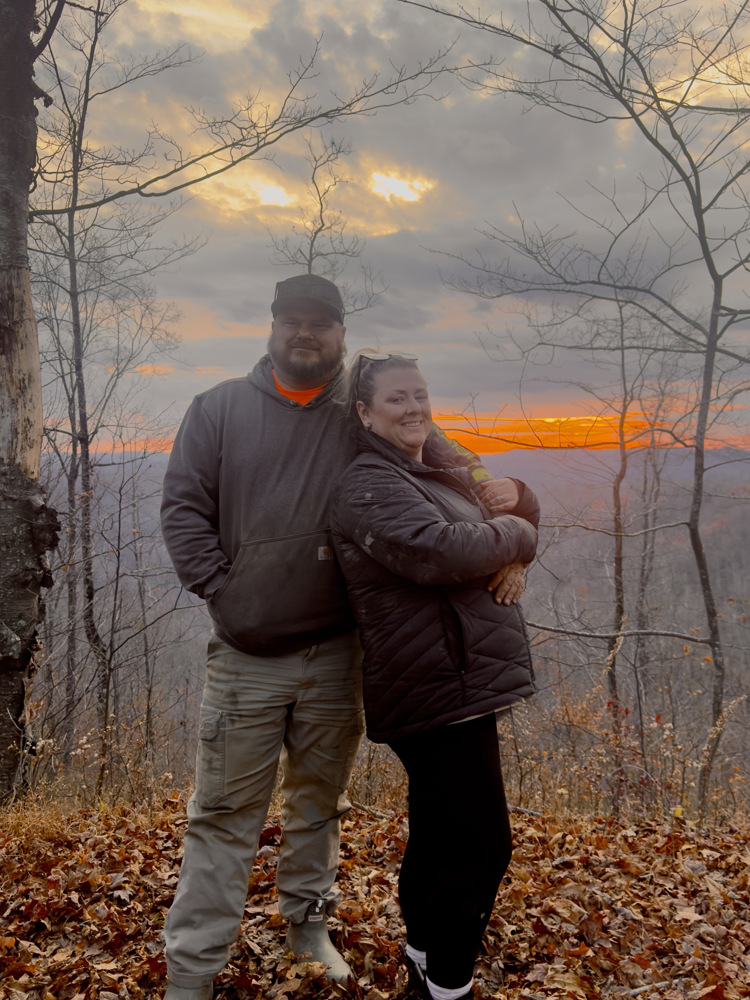 A man and woman standing close together outdoors during sunset in a forested area with leafless trees and autumn leaves on the ground.
