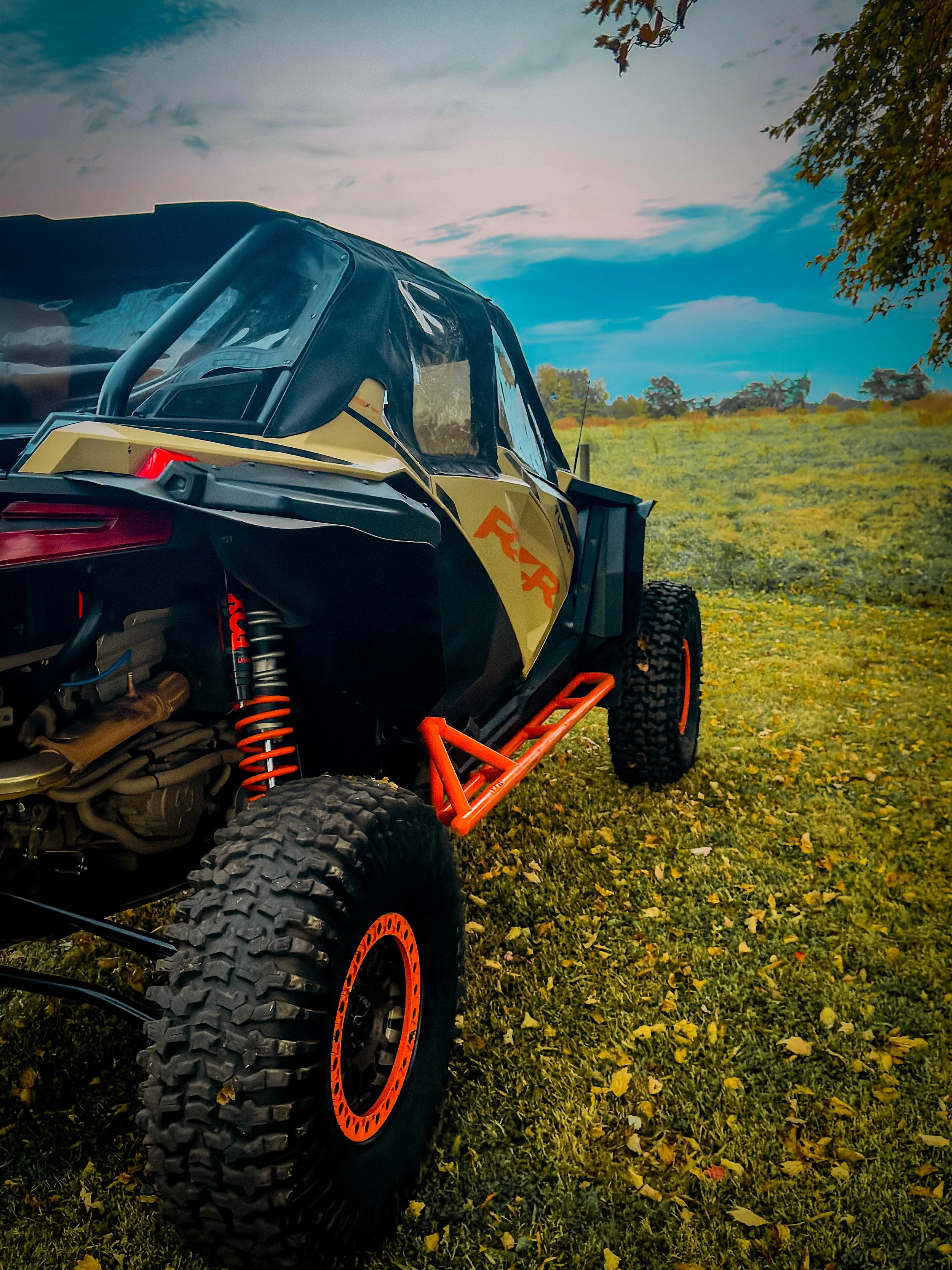 Off-road vehicle, a side-by-side ATV with black and yellow body and orange accents, parked on a grassy field with trees and a blue sky in the background.