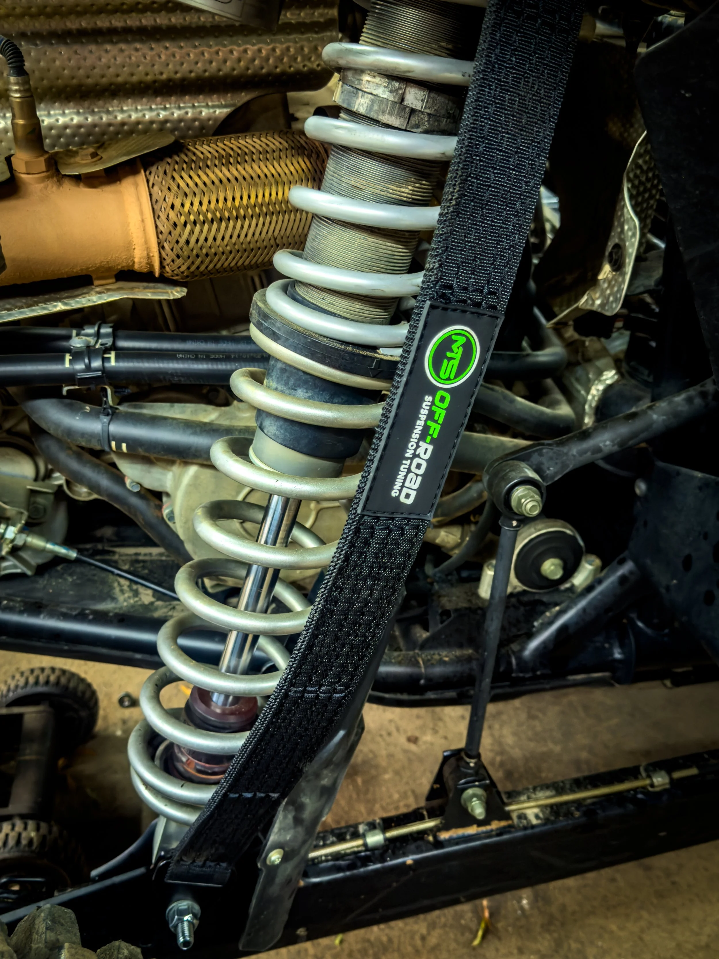 Close-up of a vehicle's suspension system featuring a coil shock absorber, with a black strap labeled 'OFF-ROAD SUSPENSION TUNING' and additional automotive components visible in the background.