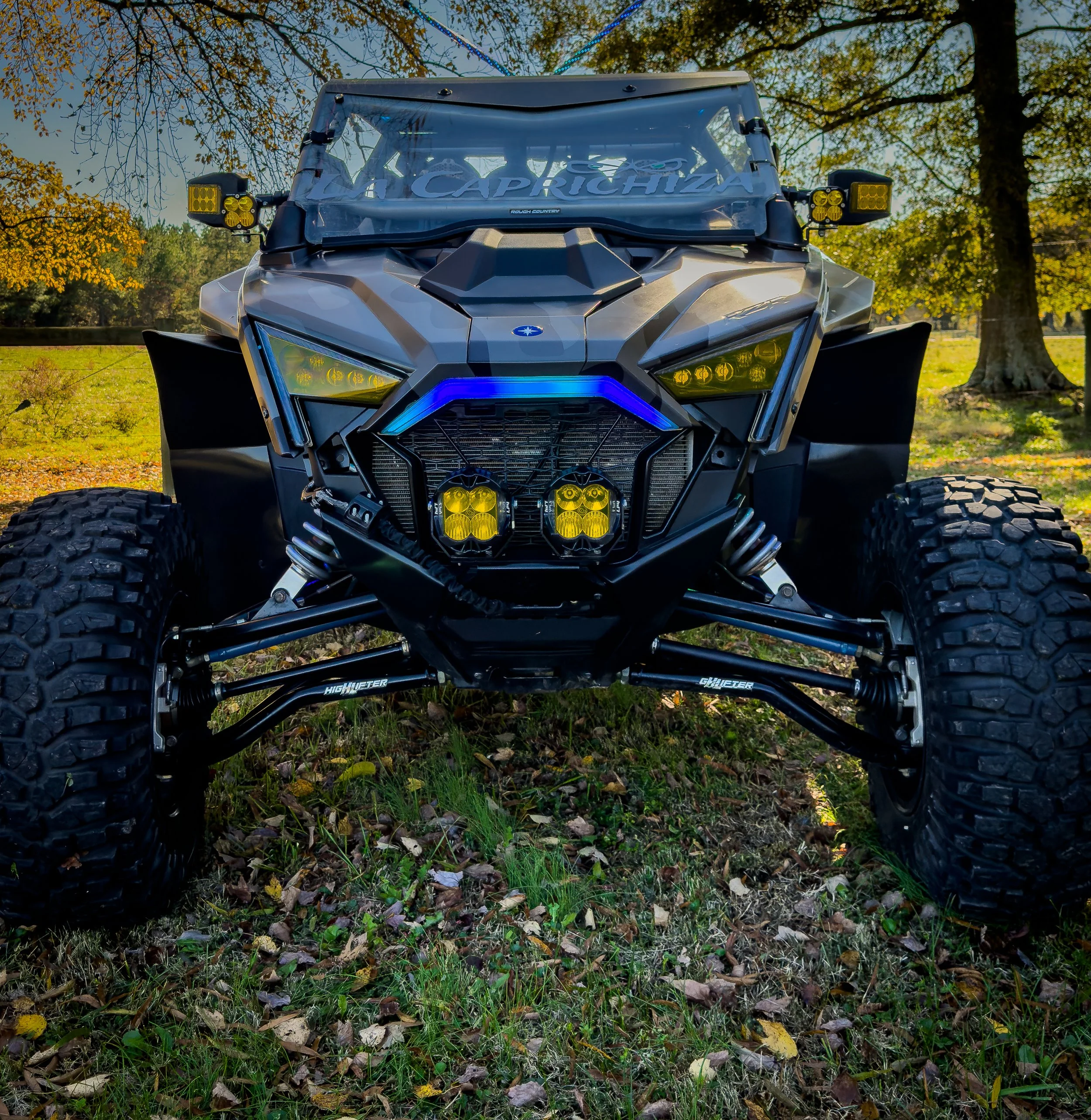 Front view of an off-road vehicle with large tires, yellow headlights, black body, and blue LED lights in a grassy field with trees and autumn leaves.