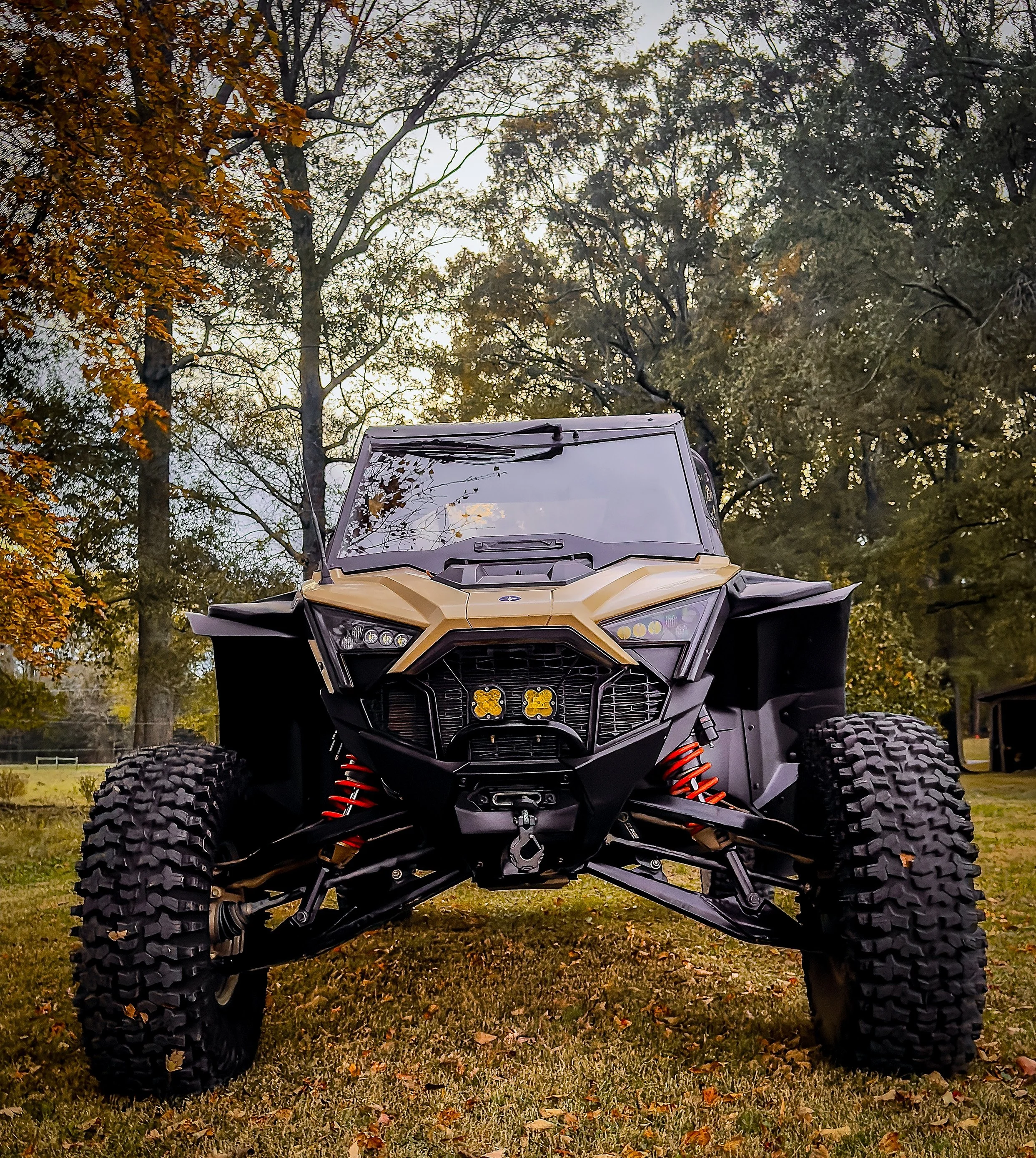An off-road vehicle, likely an ATV or UTV, parked on grass with trees in the background during fall. The vehicle has large tires, a protective front grille, and a tan and black color scheme.