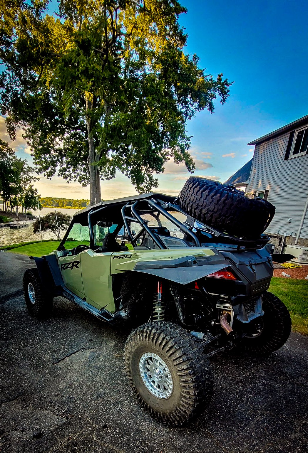 An off-road utility vehicle parked on a driveway near a tree and a house, with a large spare tire mounted on its back, water body and trees in the background, during sunset or sunrise.
