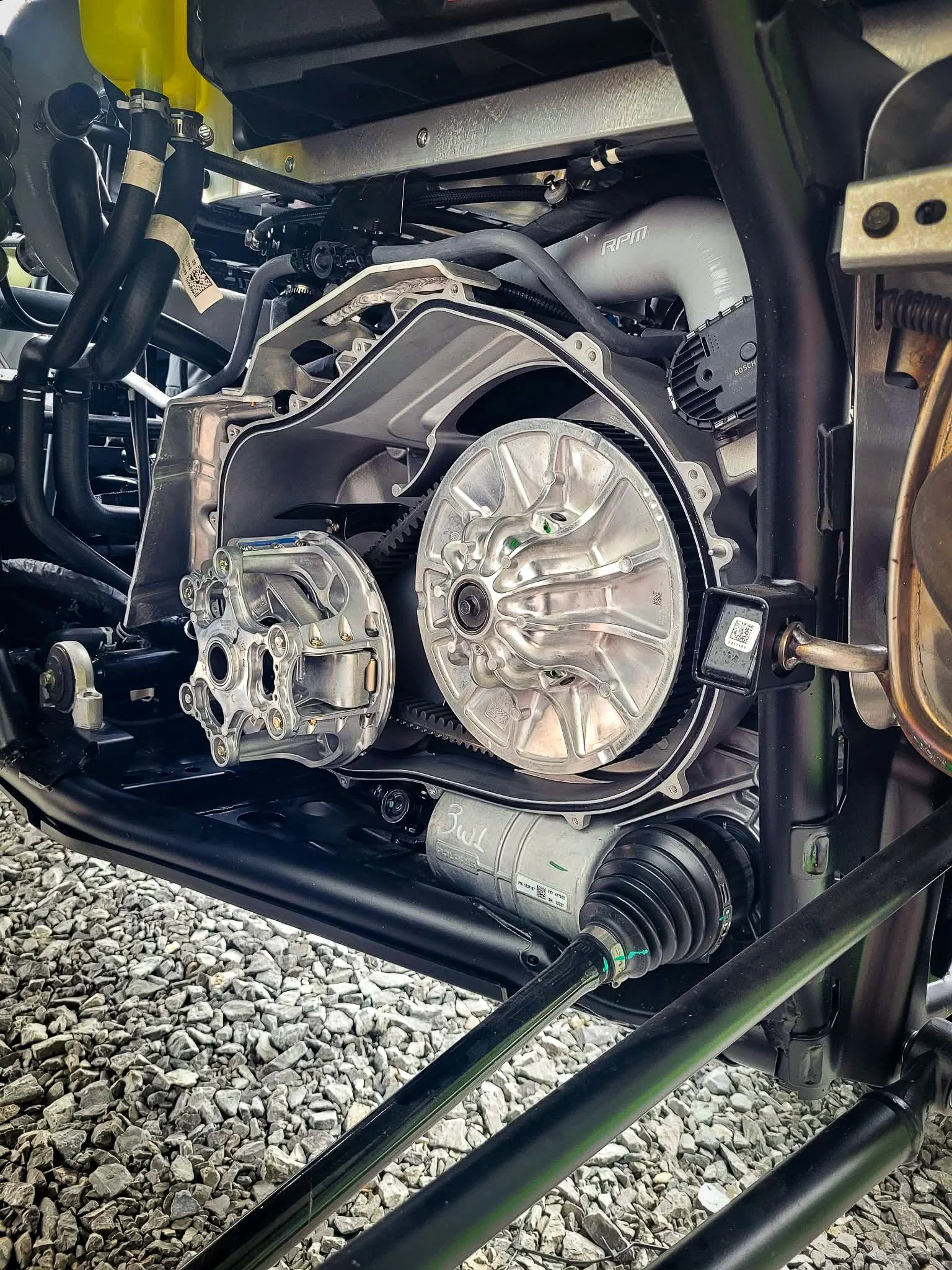 Close-up of a car's engine components, including a metallic transmission housing, belts, and a driveshaft, on a gravel surface.