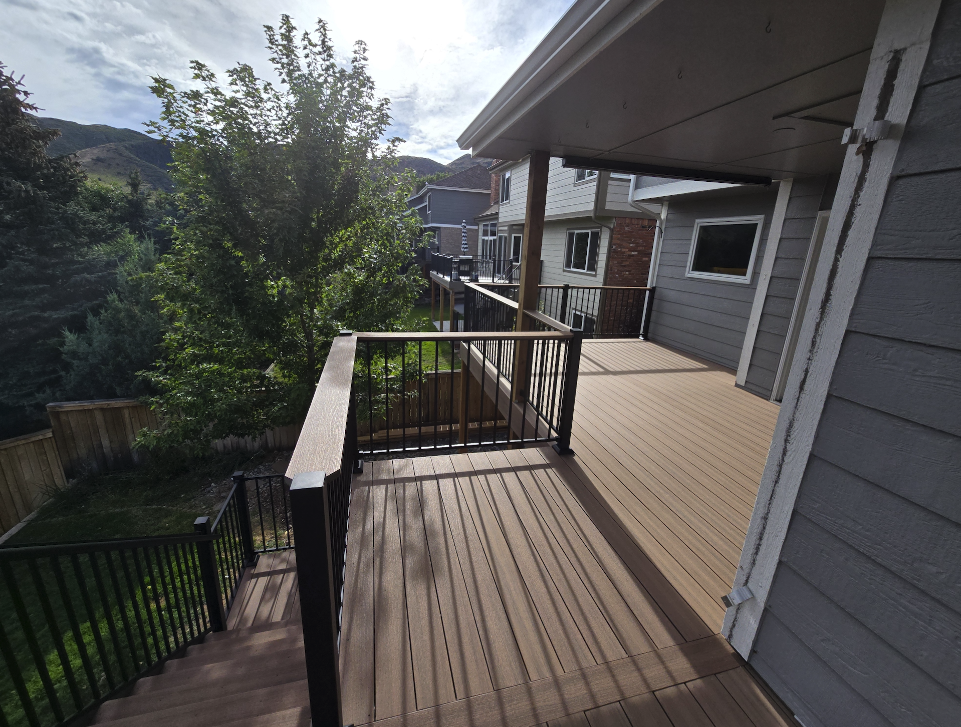 View of a deck with a built in roof cover with black metal railings attached to a house, overlooking a backyard with trees, a fence, and neighboring houses. Deckorators decking. Ken Caryl