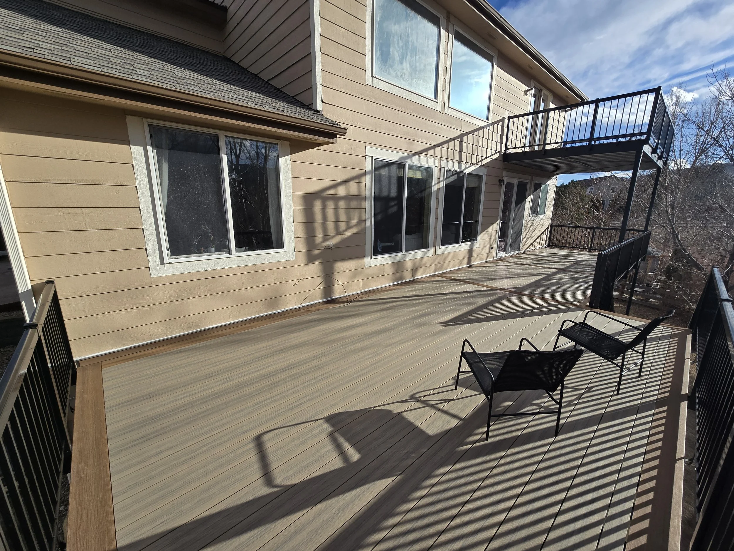 A spacious outdoor deck with a deck, two black chairs, and a railing, attached to a beige house with large windows, with a clear blue sky overhead and shadows cast by the railing. Deckorators decking. Littleton