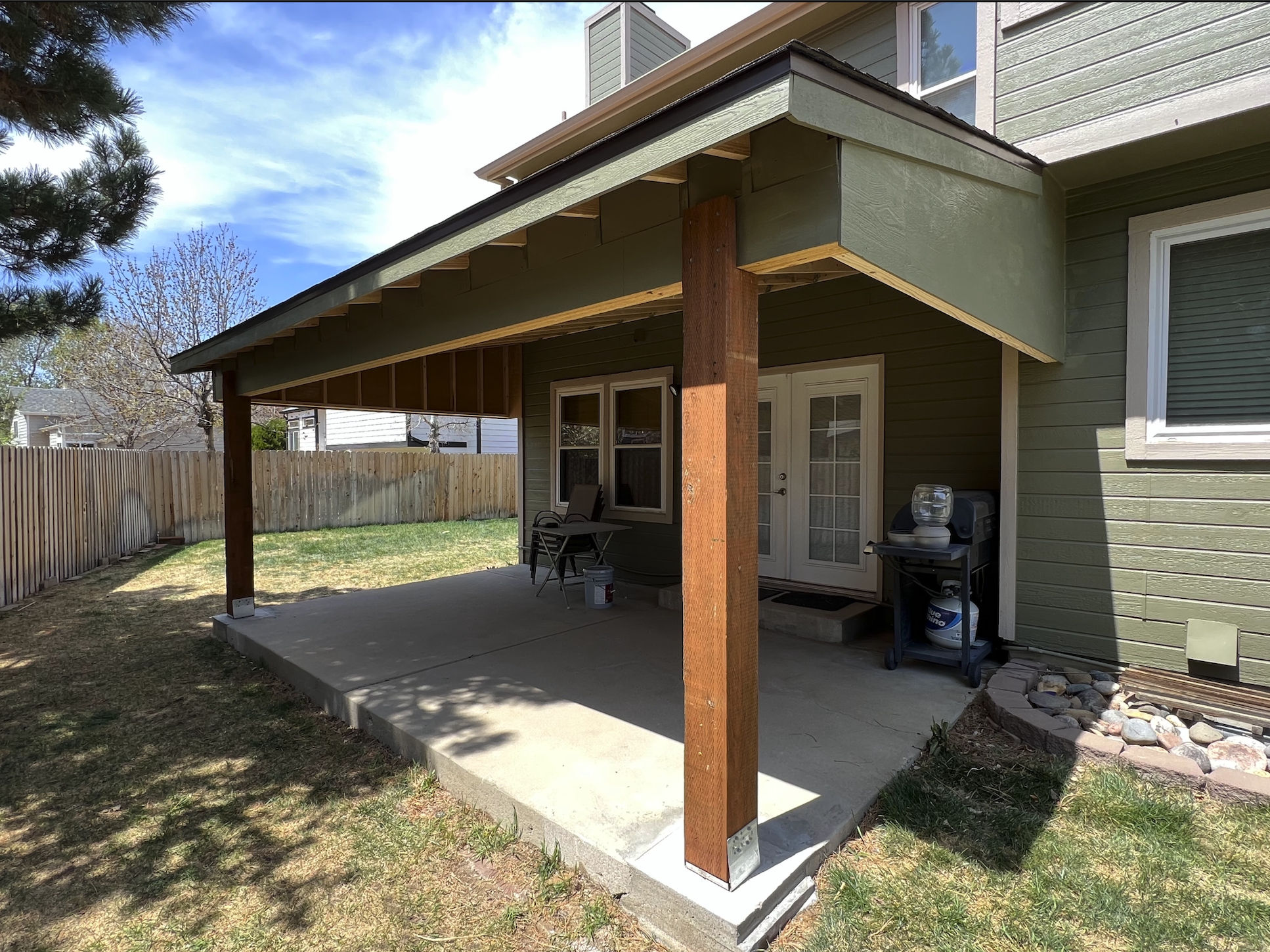 Backyard patio with a new covered porch, supported by two wooden posts, attached to a green house with white trim, featuring two windows and a door. There are chairs, a small grill, and a propane tank on the patio, with a wooden fence and some trees 