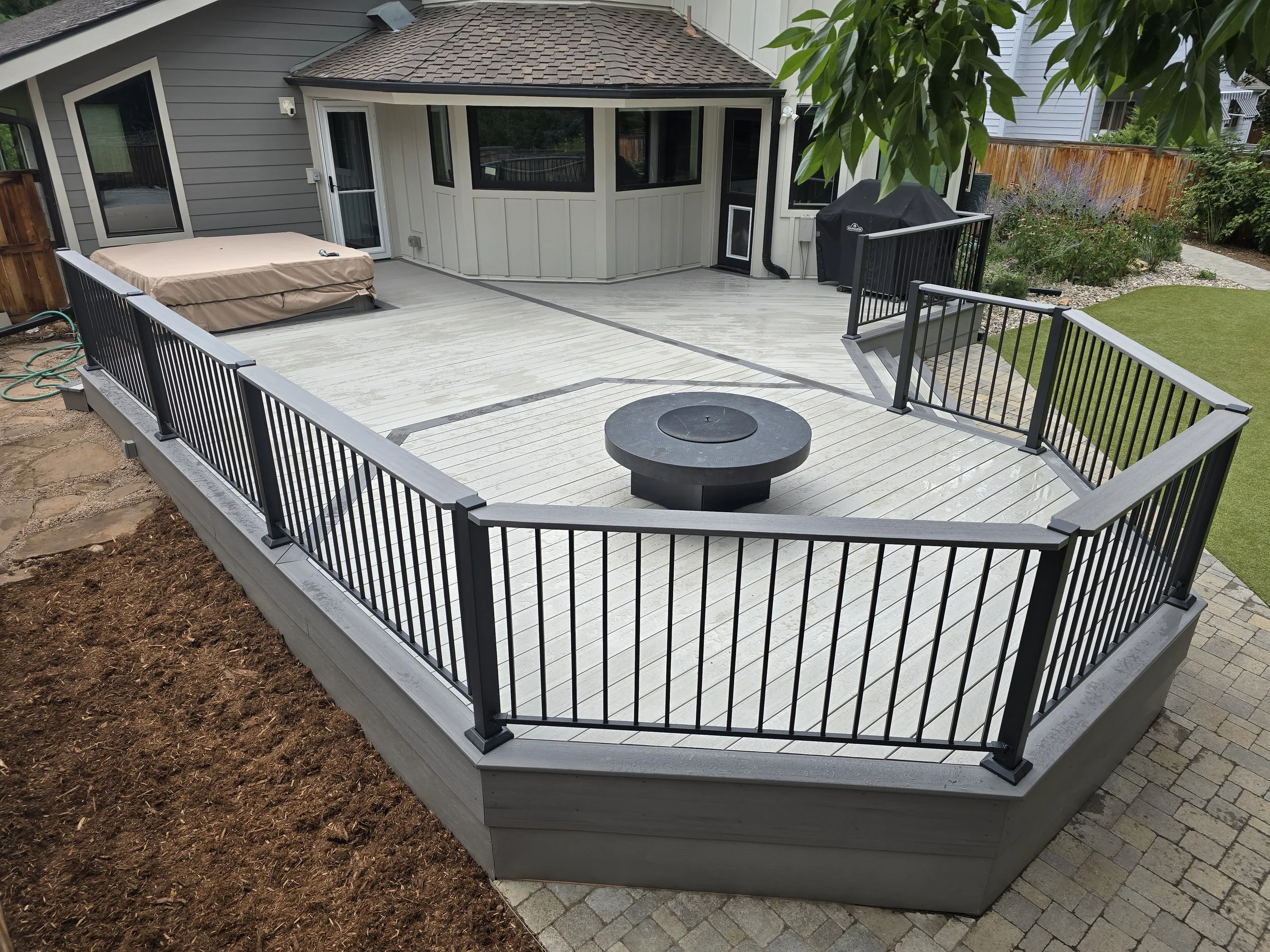 A backyard deck with black metal railings, a round fire pit in the center, and a hot tub covered with a beige cover. There is a small staircase leading down from the deck to a brick-paved patio space. The house has gray siding with a corner of a bay 