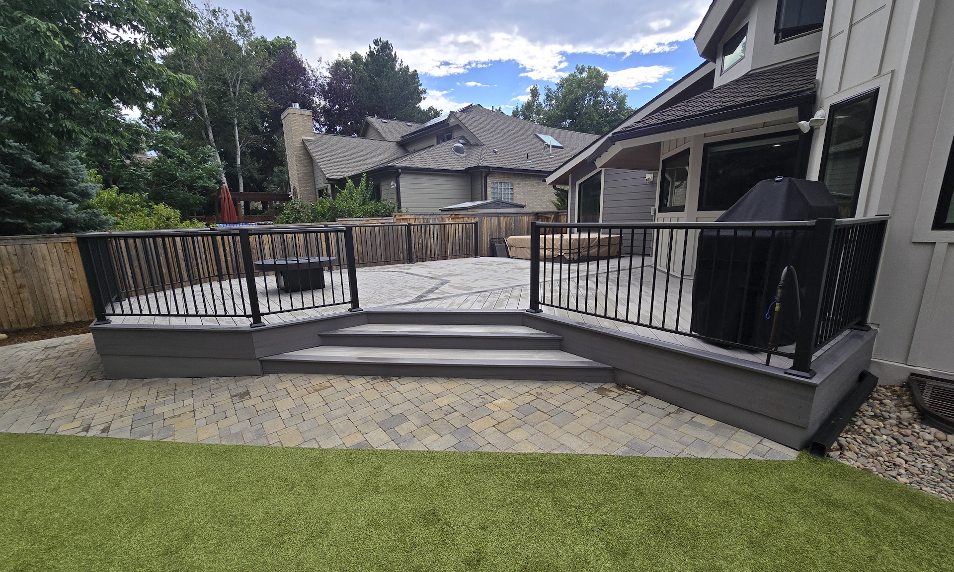 Backyard patio with steps, a black railing, a grill, outdoor furniture, and a wooden fence, with neighboring houses and trees in the background. Timbertech decking. Boulder 