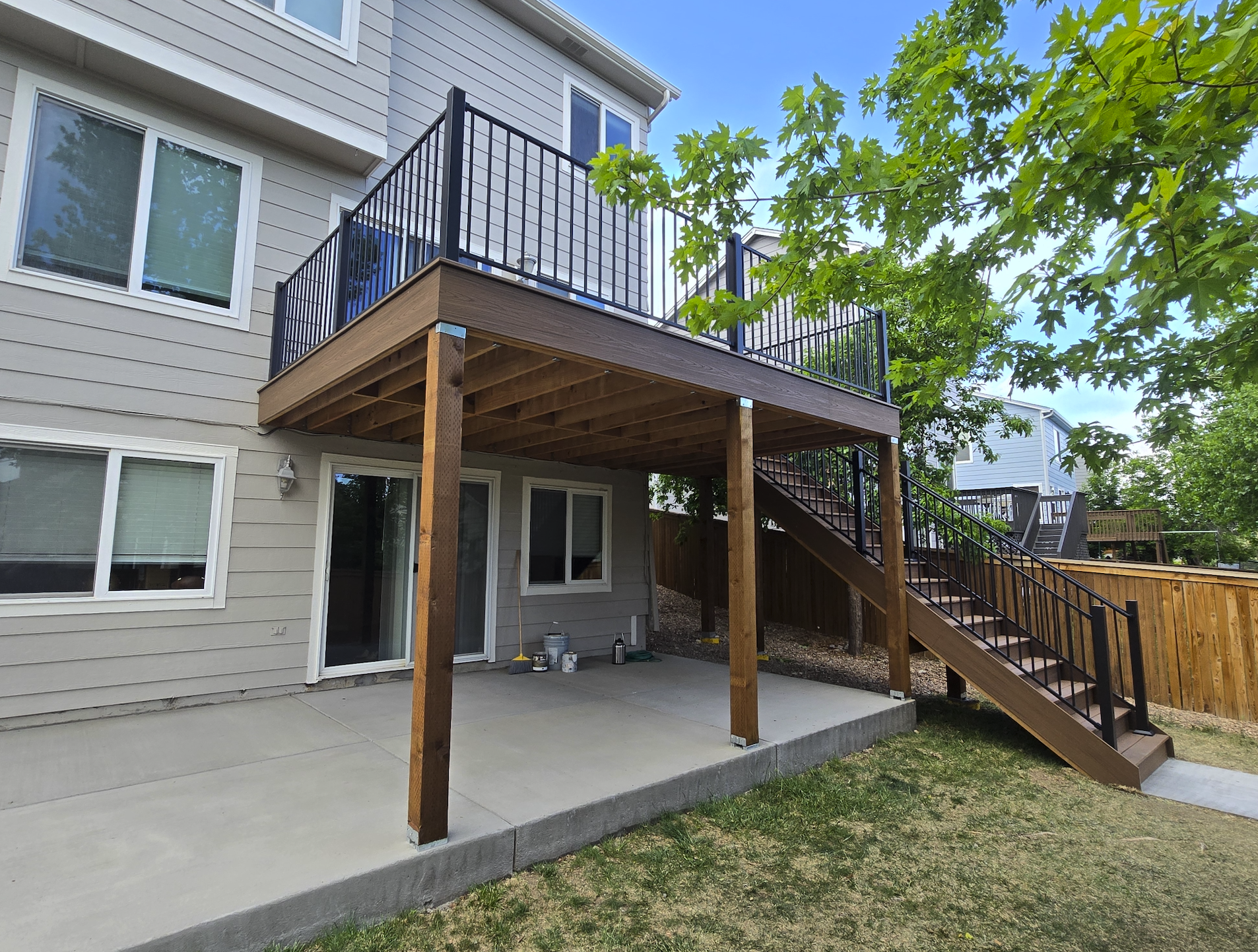 A newly built two-story house with a wooden deck extension on the ground level, supported by four wooden posts. The deck includes a staircase leading down to the yard, with a black metal railing. There are several windows and a sliding glass door on 