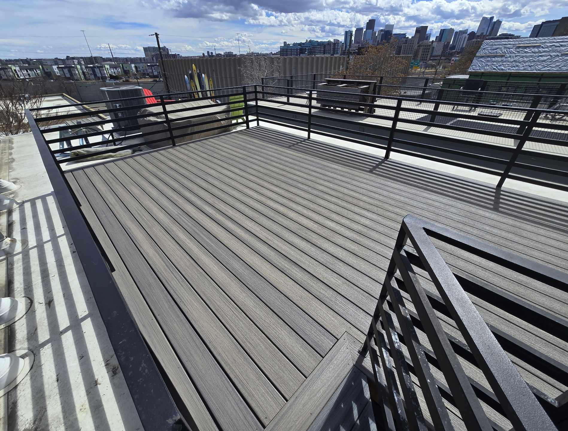 Empty rooftop deck with wooden flooring, black metal railing, and city skyline in the background under partly cloudy sky. Trex decking. 